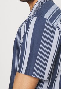 Camicia con maniche corte e bottoni, di colore blu navy con righe verticali bianche. Realizzata in tessuto leggero con una texture morbida e colletto classico.