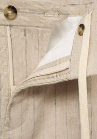 Beige gestreepte broek met een ritssluiting en knoopsluiting. Kenmerken zijn onder andere een witte voering en een geweven textuur met een trekkoord in de taille.