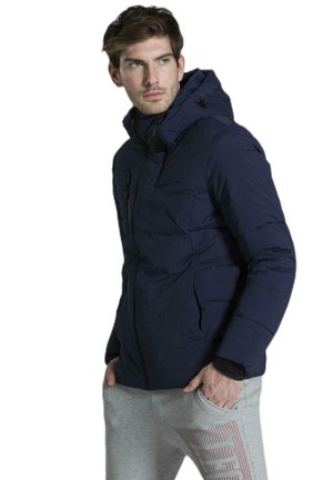 Un mannequin masculin porte une veste matelassée bleu marine à capuche et un pantalon de jogging gris. Il se tient debout, les mains dans les poches, sur un fond blanc.
