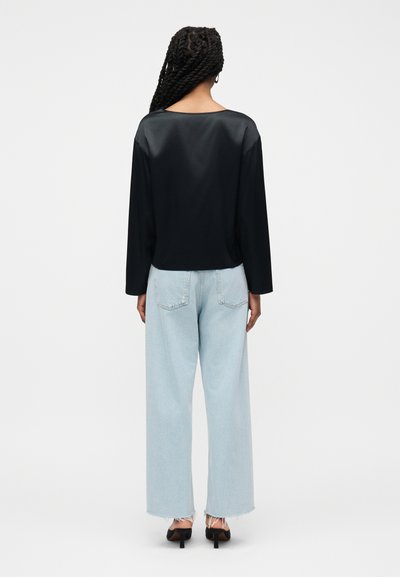 Γυναίκα με πλεγμένα μαλλιά στέκεται γυρισμένη προς τα πίσω, φορώντας μαύρο μακρυμάνικο τοπ, ανοιχτό μπλε φαρδιά τζιν παντελόνια και μαύρες γόβες.