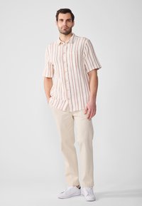 Camicia a maniche corte color crema con righe verticali rosse, con bottoni sul davanti, tasca a sinistra, abbinata a pantaloni beige e sneakers bianche.