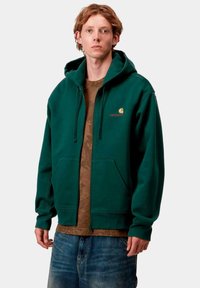 Sudadera verde con cremallera y capucha ajustable, con bolsillos tipo canguro y un acento de logo dorado. Llevada sobre una camiseta de patrón camuflaje marrón.