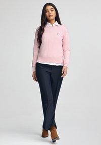 Maglione rosa chiaro con scollo rotondo e logo ricamato, abbinato a pantaloni blu scuro e stivaletti marroni. Tessuto liscio.
