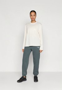 Gaiši bēša garroku krekls kopā ar tumši pelēkiem sporta biksēm ar logotipa apdruku un melniem sporta apaviem ar gludu tekstūru.