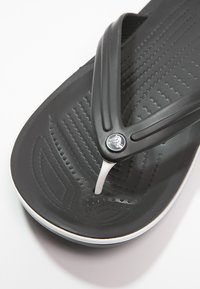 Svart flip-flop sandal med en texturerad fotbädd, vit accentrem och en logotyp på toppen. Har en vadderad sula med spår.
