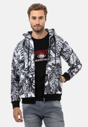 Kapuzenjacke in Schwarz und Weiß mit aufwendigen Löwen- und Tribal-Mustern. Rippstrickbündchen und Saum mit einem Reißverschluss vorne. Details an den Ärmeln.