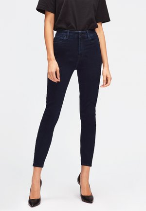 Jean skinny bleu marine en denim, avec taille haute, passants de ceinture et poches avant, assortis à des talons hauts noirs.
