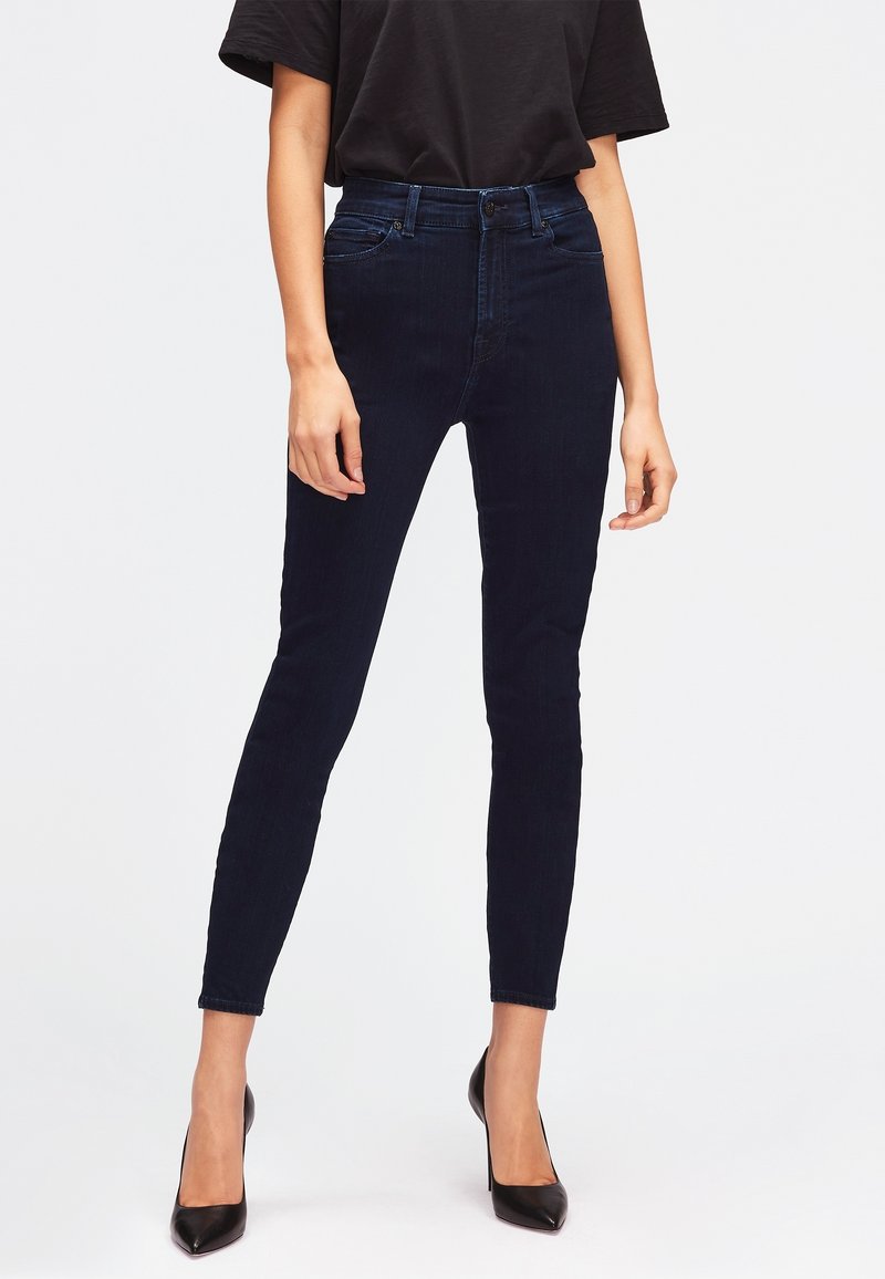 Jean skinny bleu marine en denim, avec taille haute, passants de ceinture et poches avant, assortis à des talons hauts noirs.