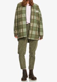 Zelená kostkovaná oversize bunda s hnědými akcenty, světle růžová košile, olivové manšestrové kalhoty a hnědé kotníkové boty. Minimalistické designové prvky.