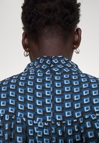 Personne aux cheveux courts et bouclés portant un haut bleu avec des motifs géométriques en carrés, montrée de dos sur un fond clair uni.