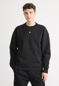 Sudadera negra hecha de tejido suave, con cuello redondo y puños y dobladillo acanalados. Un pequeño logo está centrado en la parte frontal.