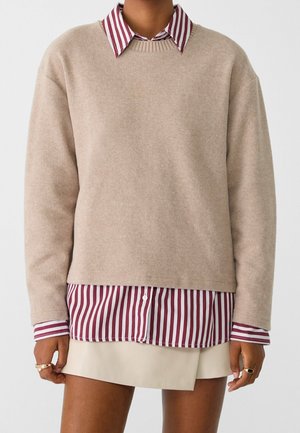 Pull beige superposé sur une chemise à rayures bordeaux et blanche, associée à une jupe mini beige, montrant des mains avec des bagues en or.