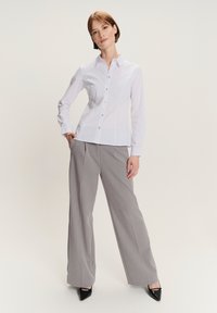 Donna in piedi con una mano in tasca, che indossa una camicia bianca con bottoni, pantaloni grigi a palazzo, tacchi neri e orecchini neri su sfondo bianco.