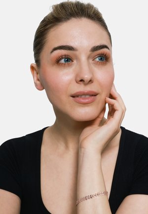 Bracelet en or rose avec un motif de feuilles porté au poignet, associé à un haut noir. La main de la personne repose près de son visage. Peau lisse, maquillage neutre.