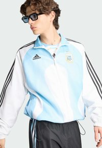 Νέος άνδρας με μπλε και λευκό μπουφάν Adidas της Αργεντινής με μαύρες ρίγες στα μανίκια και μαύρο παντελόνι, κοιτάζοντας στο πλάι με γυαλιά ηλίου.