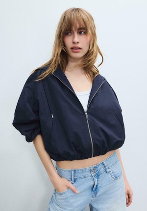 Jeune femme aux cheveux blonds portant une veste courte bleu marine zippée sur un haut blanc et un jean bleu clair, debout devant un fond uni.