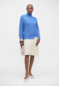 Helles blaues Strickpullover mit hohem Kragen, kombiniert mit einem cremefarbenen, knielangen Kleid. Model hält eine hellrosane Handtasche.