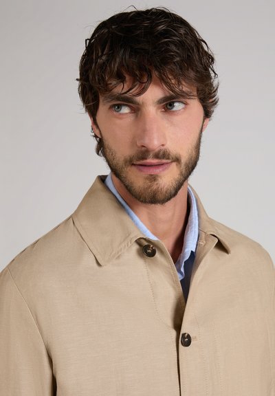 Hombre con cabello corto y rizado de color castaño y barba, vestido con un abrigo beige sobre una camisa azul claro con cuello, mirando hacia un lado sobre un fondo sencillo.