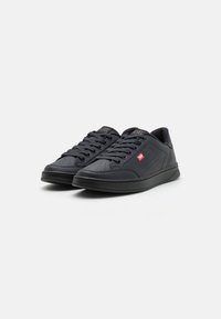 Chaussures de sport noires avec un extérieur en cuir lisse, fermeture à lacets, détails perforés et semelle en caoutchouc. Accent de logo rouge sur le côté.