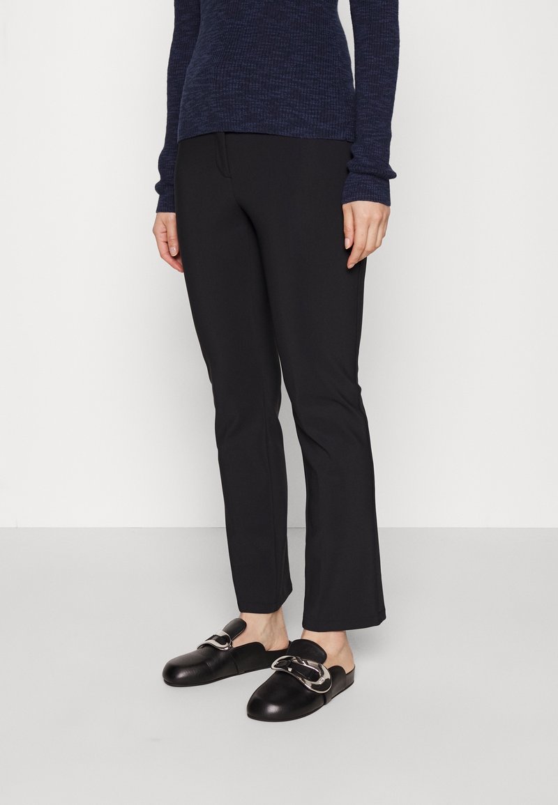 Pantalon noir ajusté avec une coupe slim, associé à des sabots noirs à bout ouvert avec un détail de boucle métallique. Pull bleu marine texturé visible.