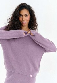 Luźny lawendowy sweter z miękkiego, puszystego materiału z długimi rękawami i bocznymi rozcięciami; ma małą różową metkę z logo przy krawędzi.