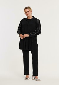 Zwarte blouse met lange mouwen en zilveren strepen, gecombineerd met zwarte broeken en metallic hakken. Soepele stof.