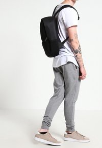 Černý batoh s hladkým povrchem a nastavitelnými popruhy, který nosí postava ve bílé tričku a šedých teplácích, stojící na neutrálním pozadí.