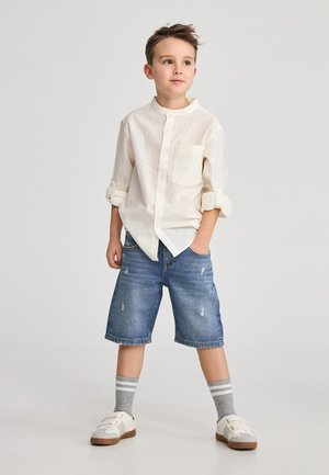 Camicia button-up beige chiaro, maniche arrotolate, piccola tasca. Pantaloni corti in denim, blu con effetto usurato. Calze grigie a righe, sneakers bianche.