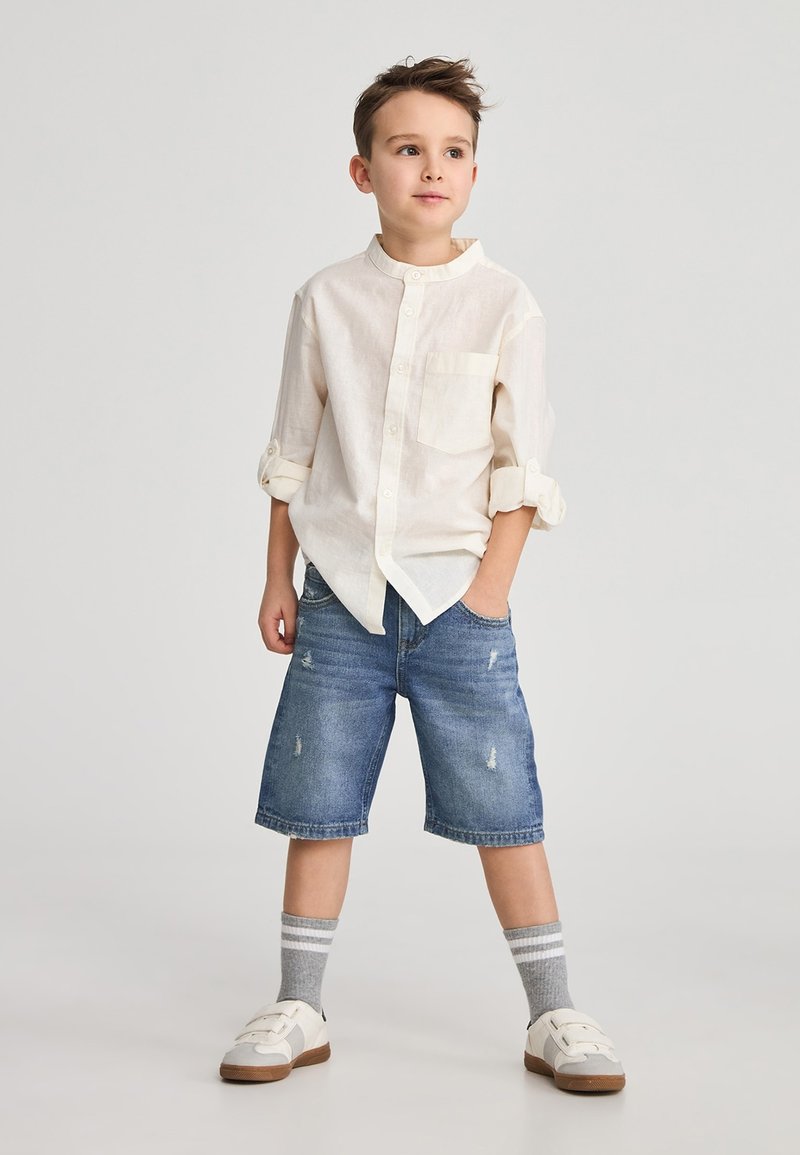 Camicia button-up beige chiaro, maniche arrotolate, piccola tasca. Pantaloni corti in denim, blu con effetto usurato. Calze grigie a righe, sneakers bianche.