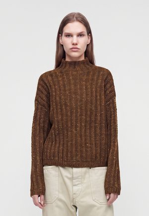Donna con lunghi capelli castani che indossa un maglione marrone a lavorazione grossa e pantaloni beige chiaro, di fronte a uno sfondo semplice.