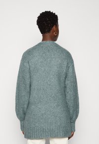 Overmaat, lange mouwen cardigan in zacht, textuurachtig teal stof met geribde zomen en manchet details. Achteraanzicht toont een ontspannen pasvorm.