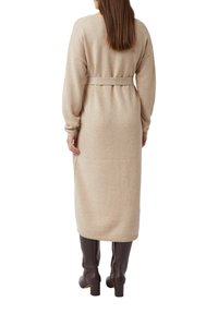 Beżowa dzianinowa sukienka z długim rękawem, z paskiem w talii i długości midi. Modelka ma na sobie brązowe kozaki za kolano na masywnym obcasie.