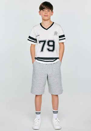 Chłopiec stoi z rękami w kieszeniach, ubrany w białą koszulkę z numerem 79, szare szorty, paskowane skarpetki i białe trampki na jednolitym tle.