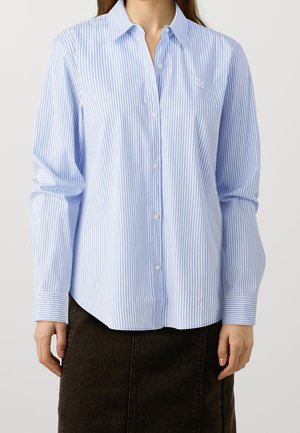 Kvinde iført en langærmet, lyseblå og hvide lodrette striped skjorte med knapper og krave samt en mørk nederdel.