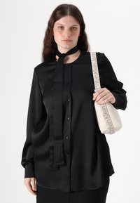 Black satin blouse with a button-down front, long sleeves, and a tied neck. Includes a cream handbag with a textured finish and patterned strap.