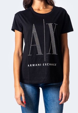 Kobieta w czarnej koszulce Armani Exchange z ćwiekowanym logo "A | X" i niebieskich dżinsach, stojąca z rozluźnionymi ramionami wzdłuż ciała.