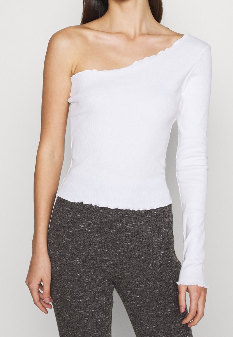 Vrouw draagt een witte, enkelschouderige top met lange mouwen in combinatie met donkergrijze geribbelde broek tegen een effen achtergrond.