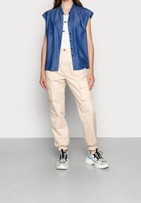 Modrá bezrukávová košile na knoflíky, béžové cargo kalhoty s elastickými manžetami a bílé tenisky s černými a modrými akcenty. Neutrální pozadí.