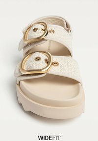 Beige slide-sandal med texturerade tygremmar, stora gyllene spännen och en tjock, kraftig sula. Accenter inkluderar metalliska detaljer.