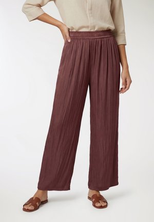 Donna con pantaloni larghi a vita alta color malva stropicciati con vita elastica, top beige e sandali slip-on marroni.