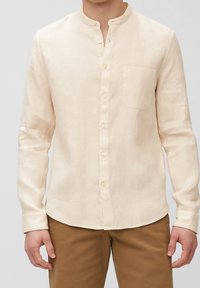 Uomo che indossa una camicia a maniche lunghe color beige con colletto alla mandarino, bottoni frontali e taschino sul petto, abbinata a pantaloni marroni.