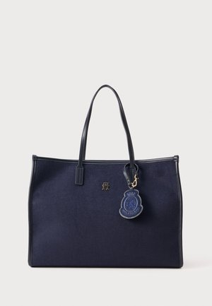 Navy fabric tote bag with two leather handles, gold logo emblem, and a blue logo charm attached with a small clasp detail.