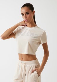 Beige lyhyt t-paita, joka on valmistettu pehmeästä materiaalista ja jossa on "GUESS"-logo strasseilla. Malli poseeraa yksi käsi kaulalla vaaleaa taustaa vasten.