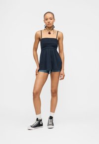 Νέα γυναίκα με πλεξούδες, φορώντας σκούρο μπλε αμάνικο τοπ, τζιν σορτς, λευκές κάλτσες και μαύρα ψηλά αθλητικά παπούτσια, στέκεται μπροστά σε λευκό φόντο.