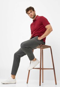 Polo bordeaux avec rayures blanches, pantalon gris ajusté et baskets blanches. Modèle assis sur un tabouret en cuivre contre un fond uni.