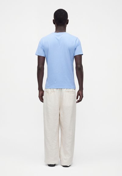 Homme debout de dos, portant une chemise bleu clair à manches courtes et un pantalon blanc ample sur un fond uni.