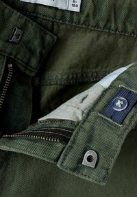 Pantalon en coton vert avec une fermeture éclair, quincaillerie en métal et étiquettes en tissu. Présente un tissu intérieur contrastant et un bouton à rabat.