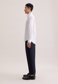 Biała koszula z długim rękawem o subtelnym wzorze, zestawiona z granatowymi spodniami o szerokich nogawkach oraz czarnymi eleganckimi butami. Widok z profilu.