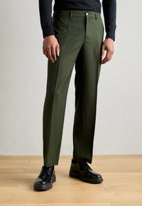 Uomo che indossa pantaloni su misura verde oliva con piega, camicia nera a maniche lunghe e scarpe eleganti lacciate nere, in piedi su un pavimento in legno.