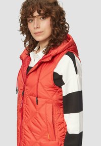 Roter gesteppter Weste mit Kapuze, ausgestattet mit verstellbaren Zugbanden, Seitentasche und einem kontrastierenden schwarz-weiß gestreiften Langarmshirt darunter.